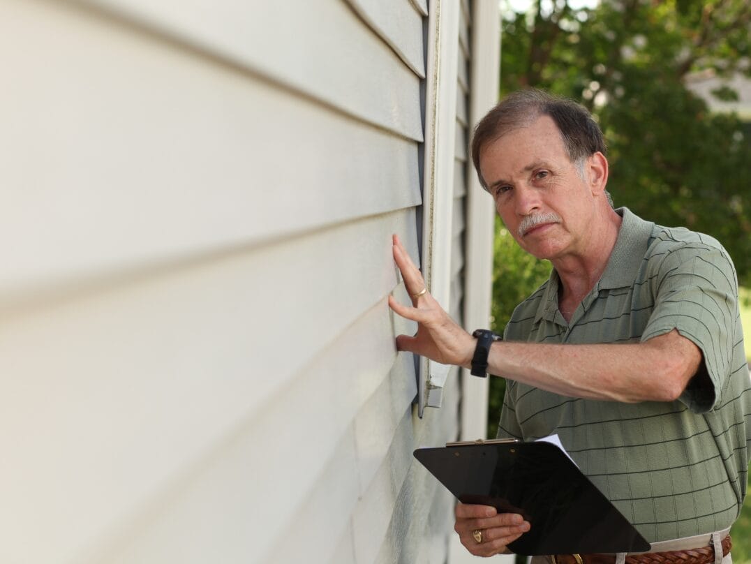 Home inspector examining siding of a house while holding a clipboard